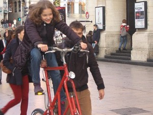 Jeune personne sur un tall-bike rouge, se faisant assurée par un-e adulte