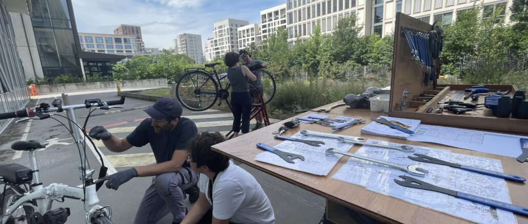 La remorque atelier-mobile est déployée. On voit des outils et deux personnes entrain de réparer un vélo